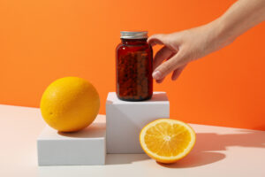 Hangi Vitamin Eksikliği Baş Dönmesi Yapar? 2 Portakal ve limon yanında, elin tuttuğu amber renkli vitamin şişesi; turuncu arka plan üzerinde C vitamini konseptli ürün görseli.